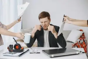 Adult man stressing out at home office desk with multiple demands surrounding him.