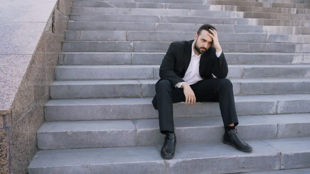 A businessman in a suit sits on stone steps, looking stressed and thoughtful outdoors.