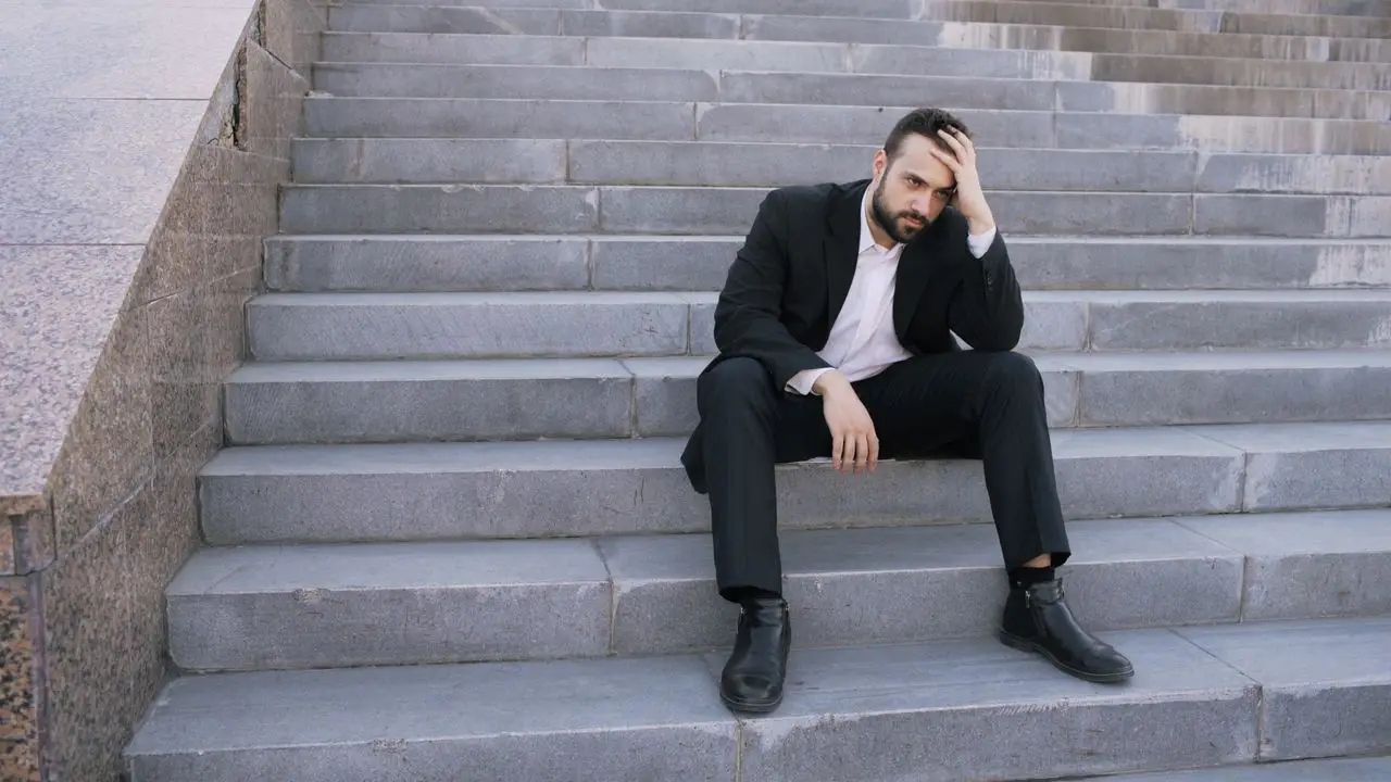 A businessman in a suit sits on stone steps, looking stressed and thoughtful outdoors.