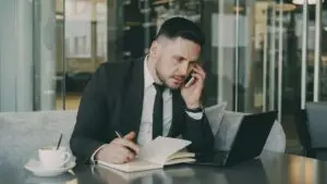 Businessman talks on phone while writing notes in a modern cafe.